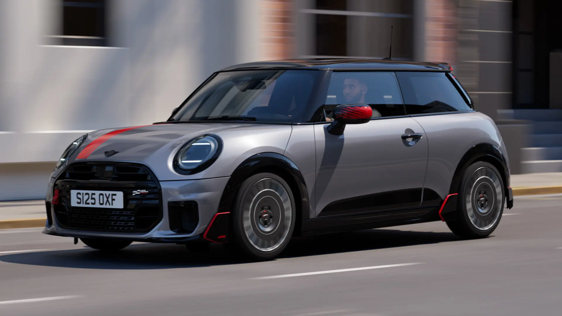 MINI Cooper 3-door in Melting Silver with John Cooper Works mirror cap and bonnet decal in Chili Red, roof spoiler and winglet in Black, driving along city street – front-side view.